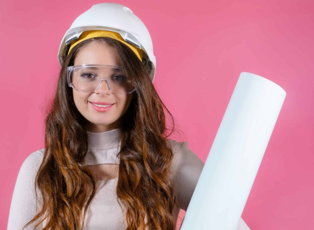 Young female architect in helmet and goggles, holding her project exuding confidence and enthusiasm